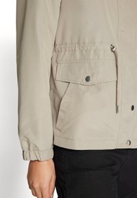 Lys beige zip-jakke med elastiske mansjetter, innsnørt midje og frontlomme. Glatt stoff med subtil glans og sølvfargede detaljer.