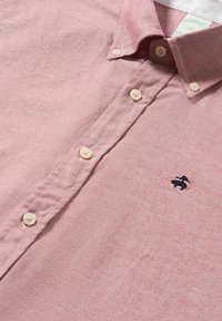 Světle růžová button-up košile vyrobená z bavlny, s bílým límcem, sedmi knoflíky a malým černým vyšitým logem na levé hrudi.