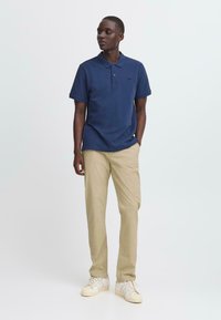 Navy polo shirt made of cotton, short sleeves, with a relaxed fit. Paired with light beige pants and white sneakers, standing against a plain background.