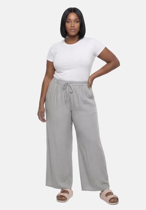 Kobieta stoi z ręką na biodrze, ubrana w białą koszulkę z krótkim rękawem, jasnoszare szerokie spodnie ze sznurkiem i beżowe sandały.