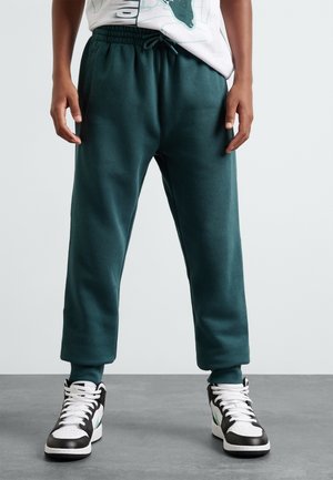 Niño con pantalones deportivos verde oscuro y zapatillas blanco y negro, de pie sobre un suelo gris con fondo de pared blanca.