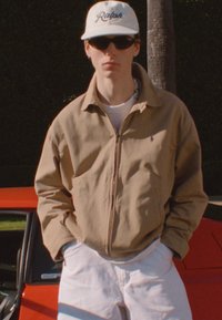 Young person wearing beige jacket, white cap with "Ralph," sunglasses, and white pants stands in front of a red car outdoors.