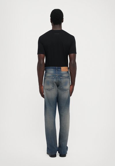 Man som står med ryggen mot oss, klädd i en svart kortärmad skjorta och blekta blå jeans med bakfickor och läderdetaljer.