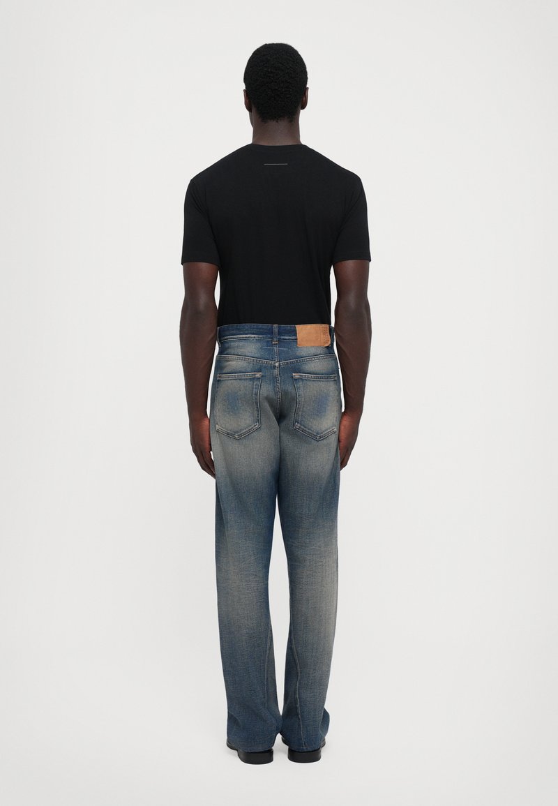 Man som står med ryggen mot oss, klädd i en svart kortärmad skjorta och blekta blå jeans med bakfickor och läderdetaljer.