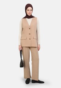Pantaloni beige e un gilet abbinato con bottoni frontali, abbinati a una maglietta bianca a maniche lunghe e un hijab marrone scuro, oltre a scarpe nere e una borsa.