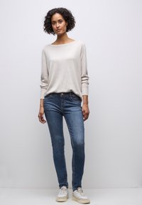 Camisa de manga larga color beige claro combinada con jeans ajustados azul oscuro y zapatillas blancas. Textura suave, diseño casual y ajuste holgado.