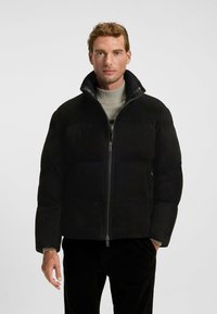Sort pufferjakke med høj krave, glat tekstur, lynlås foran og paneldesign. Båret over en lys grå sweater.