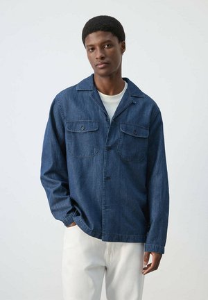 Jonge man die een blauwe spijkerblouse met knopen draagt over een wit t-shirt en witte broek, staand tegen een effen lichtgrijze achtergrond.