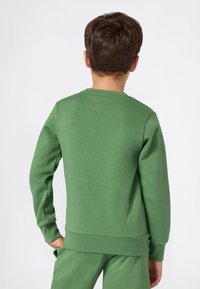 Kind in een groene sweatshirt met lange mouwen en bijpassende broek, van achteren gezien op een effen witte achtergrond.