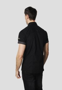 Camisa negra de manga corta con cuello abotonado, con detalles en contraste en los puños y una textura suave. Diseño simple, sin patrones visibles.