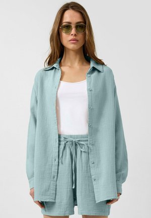 Kobieta ubrana w jasnoniebieską teksturowaną koszulę na guziki i pasujące krótkie spodenki, na białym topie, z okularami przeciwsłonecznymi o zielonym odcieniu, zwrócona przodem.