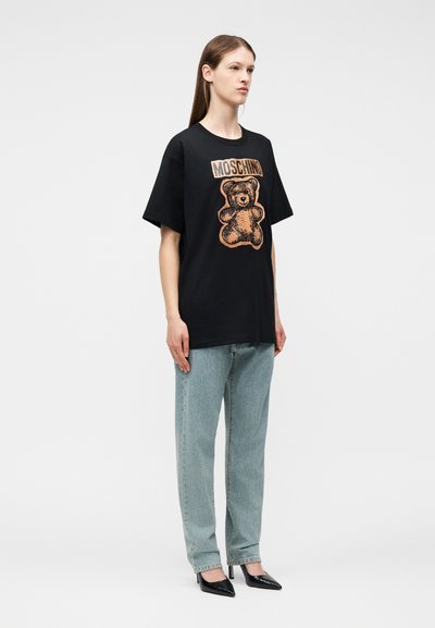 Γυναίκα που φοράει μαύρο t-shirt Moschino με εκτύπωση αρκουδάκι, ανοιχτόχρωμα τζιν και μαύρα μυτερά τακούνια, στέκεται μπροστά σε απλό φόντο.