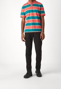 Polokošile s růžovými a tyrkysovými vodorovnými pruhy, krátké rukávy a límec; v kombinaci s černými kalhotami typu cargo a černými teniskami.