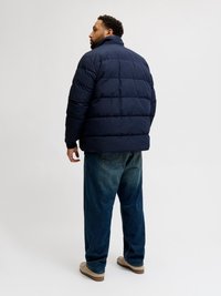 Giacca puffer blu navy con colletto alto e design trapuntato, abbinata a jeans in denim blu larghi e scarpe di colore chiaro.