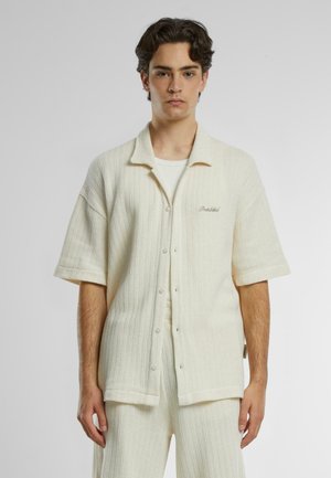 Camisa de color crema, de manga corta, con un diseño texturizado y acanalado. Presenta un cuello, botones y un logotipo bordado en el lado izquierdo.