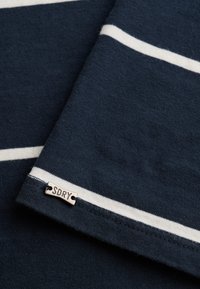 Mørkeblåt stof med hvide vandrette striber og en lille metaltag mærket "SDRY" syet nær kanten.