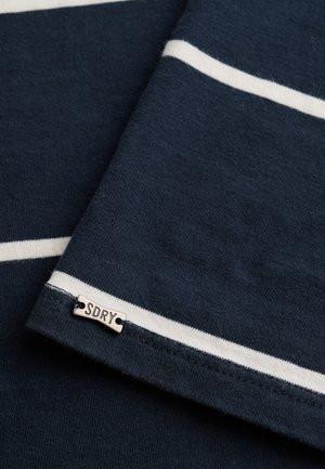 Tela azul oscuro con rayas horizontales blancas y una pequeña etiqueta de metal con la inscripción "SDRY" cosida cerca del borde.