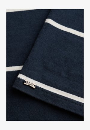 Tela azul oscuro con rayas horizontales blancas y una pequeña etiqueta de metal con la inscripción "SDRY" cosida cerca del borde.