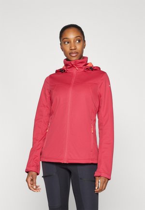 Eine Frau in einer roten Zip-Jacke mit Kapuze und schwarzen Leggings steht vor einem schlichten, hellen Hintergrund und blickt zur Seite.