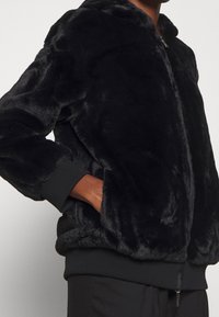 Zwarte faux fur jas met een capuchon, elastische manchetten en een ritssluiting aan de voorkant. Zachte textuur met een licht glanzende afwerking.