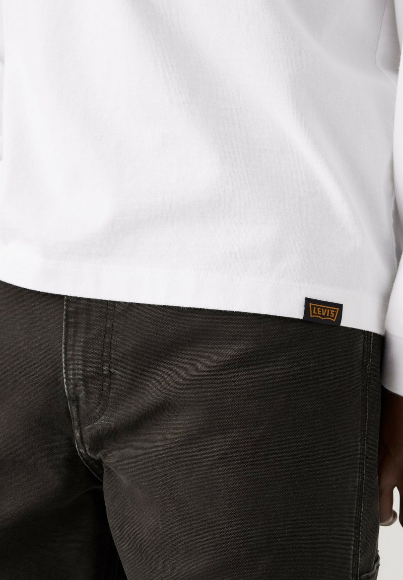 Close-up af en person iført mørke bukser og en hvid skjorte med en lille Levi's logo-tag på skjortens kant.