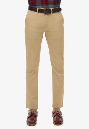 Chinos caqui de corte slim, confeccionados en suave tela de algodón. Presentan bolsillos delanteros, un cinturón de cuero marrón y zapatos de barco marrones.