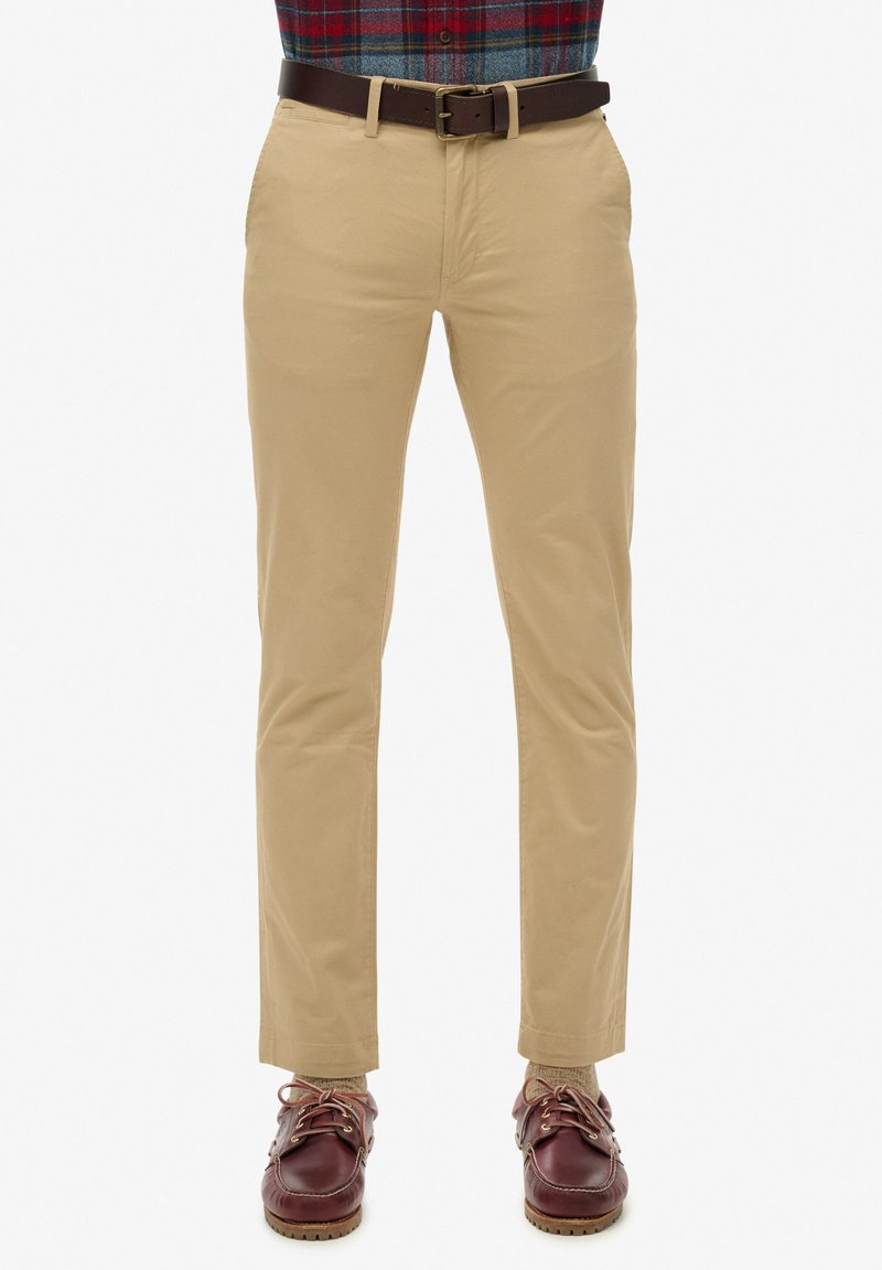 Chinos khaki con una vestibilità slim, realizzati in tessuto di cotone liscio. Presentano tasche frontali, una cintura in pelle marrone e scarpe da barca marroni.