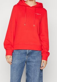 Sudadera de algodón roja con un bolsillo tipo canguro, cordones ajustables y puños acanalados, combinada con jeans de mezclilla azul claro. Detalle del logo en blanco en el pecho.
