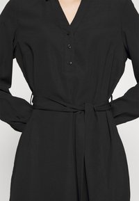 Robe noire avec encolure en V, patte de boutonnage, manches longues et ceinture assortie en tissu. Fabriquée en matériau lisse et léger.