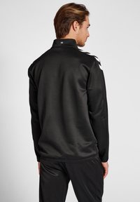 Zwarte sportjack met een staande kraag, gladde textuur en witte zijstrepen. Heeft een klein logo op de rug nabij de kraag.