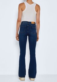 Tmavě modré zvonové džíny s vysokým pasem, zadními kapsami a hnědým koženým štítkem na pase. Nosené s bílým tílkem.