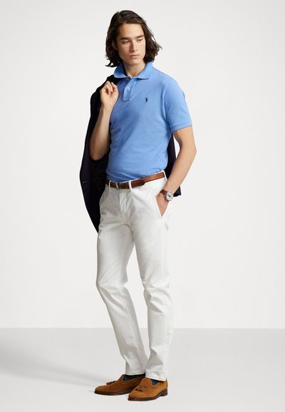 Polo bleu clair à manches courtes avec un petit logo, pantalon blanc, ceinture marron et mocassins marron. Blazer noir drapé sur l'épaule.