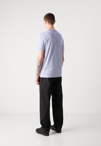 Camiseta de manga corta color púrpura claro con cuello redondo, combinada con pantalones negros holgados. Detalle de tatuaje visible en el brazo izquierdo.