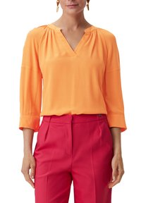 Helles orangefarbenes V-Ausschnitt-Bluse mit Dreiviertelärmeln, glatter Textur und dezentem Gathering an den Schultern, kombiniert mit hoch taillierten rosa Hosen.