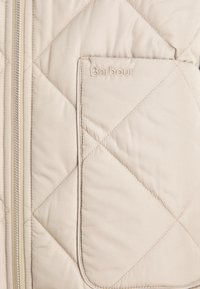 Μπεζ επενδεδυμένο μπουφάν με φερμουάρ, διαθέτοντας τσέπη στο στήθος και ανάγλυφο λογότυπο στην τσέπη. Μαλακή υφή.