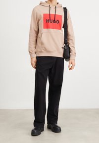 Sudadera beige con un logo rectangular rojo que dice "HUGO", pantalones negros y botas negras gruesas. La sudadera tiene un bolsillo frontal y cordones.