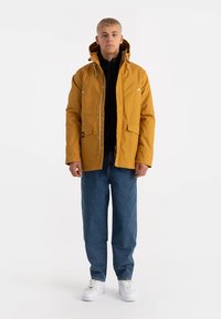 Mosterdgele buitenjacket met een capuchon, ontworpen met drukknoopsluitingen en twee voorzakken. Draagt over een zwarte hoodie en blauwe jeans.