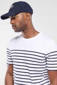 Mand iført en navy-blå cap og hvid t-shirt med lodrette navy-striber, som kigger nedad mod en ensfarvet lys baggrund.