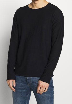 Chemise noire à manches longues en tissu doux et extensible, avec un col arrondi et de légentes fentes sur les côtés à l'ourlet pour faciliter le mouvement.