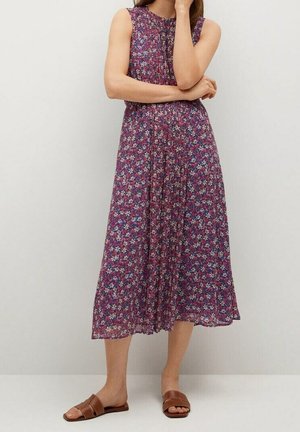Femme portant une robe midi sans manches à fleurs violettes avec des plis et des sandales marron à enfiler, debout contre un mur clair uni.