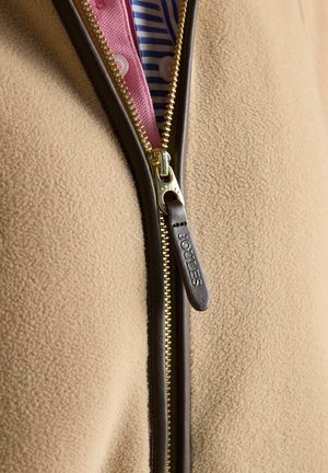 Bēša flīsa jaka ar brūnu ādas apdari zelta rāvējslēdzējam, daļēji atvērta, atklājot zem tās rozā un zilas svītrainas krekliņu.