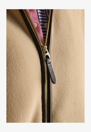 Bēša flīsa jaka ar brūnu ādas apdari zelta rāvējslēdzējam, daļēji atvērta, atklājot zem tās rozā un zilas svītrainas krekliņu.