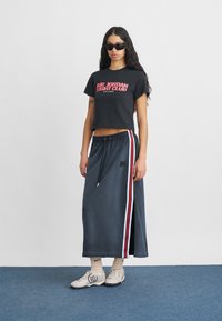 Γυναίκα με μαύρο crop top "Air Jordan Flight Club", μακριά γκρι φούστα με κόκκινες και λευκές ρίγες, λευκές κάλτσες και γκρι αθλητικά παπούτσια, στέκεται μέσα σε κλειστό χώρο.