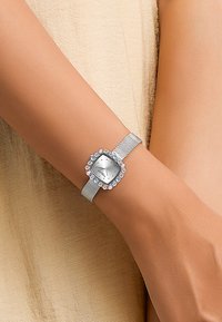 Montre-bracelet en maille argentée avec un cadran carré, un fond gris et des accents en cristal rose et bleu autour de la lunette.