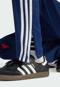 Pantalones de chándal en color navy con rayas blancas, con una cremallera lateral. Zapatillas Adidas Samba negras con rayas blancas y letras doradas en el lado.