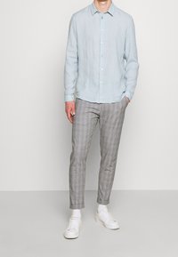 Uomo che indossa una camicia button-up azzurro chiaro, pantaloni slim-fit grigio a quadri, calze bianche e sneaker bianche, in piedi con una mano in tasca.