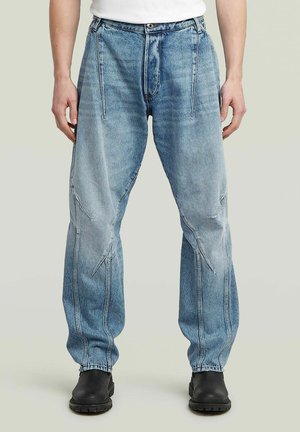 Hellblaue Denim-Jeans mit lockerer Passform, auffälligen Nähten und vorderen Falten. Mit Knopfverschluss und rohen Säumen. Kombiniert mit schwarzen Stiefeln.