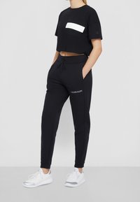 Zwarte cropped t-shirt met een witte rechthoekige patch pocket, gecombineerd met zwarte joggingbroek met het "CALVIN KLEIN" logo in het wit. Witte sneakers.