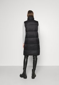 Schwarze Steppweste mit hohem Kragen, gestepptem Design und einem langen, geraden Schnitt. Getragen über einem grauen Pullover und kombiniert mit schwarzen Lederhosen.