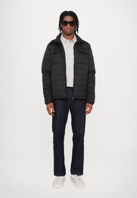 Giubbotto imbottito nero con zip frontale, due tasche sul petto, polo grigia, jeans scuri e sneakers bianche. Il modello indossa occhiali da sole scuri.